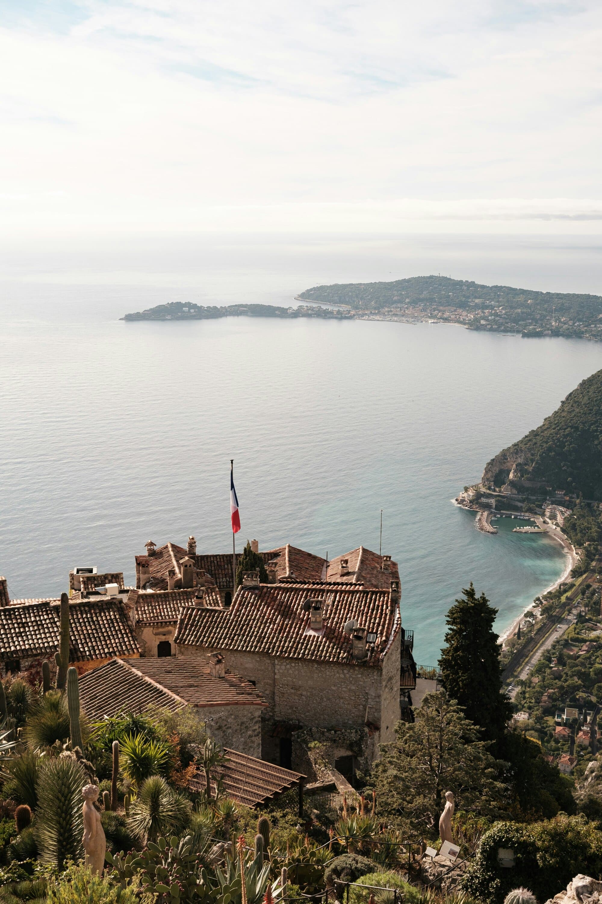 Èze rooftops with the French flag and the Mediterranean below, Saint-Jean-Cap-Ferrat in the bay — the medieval village used by The Transporter and the cinematic vantage above Monte-Carlo.